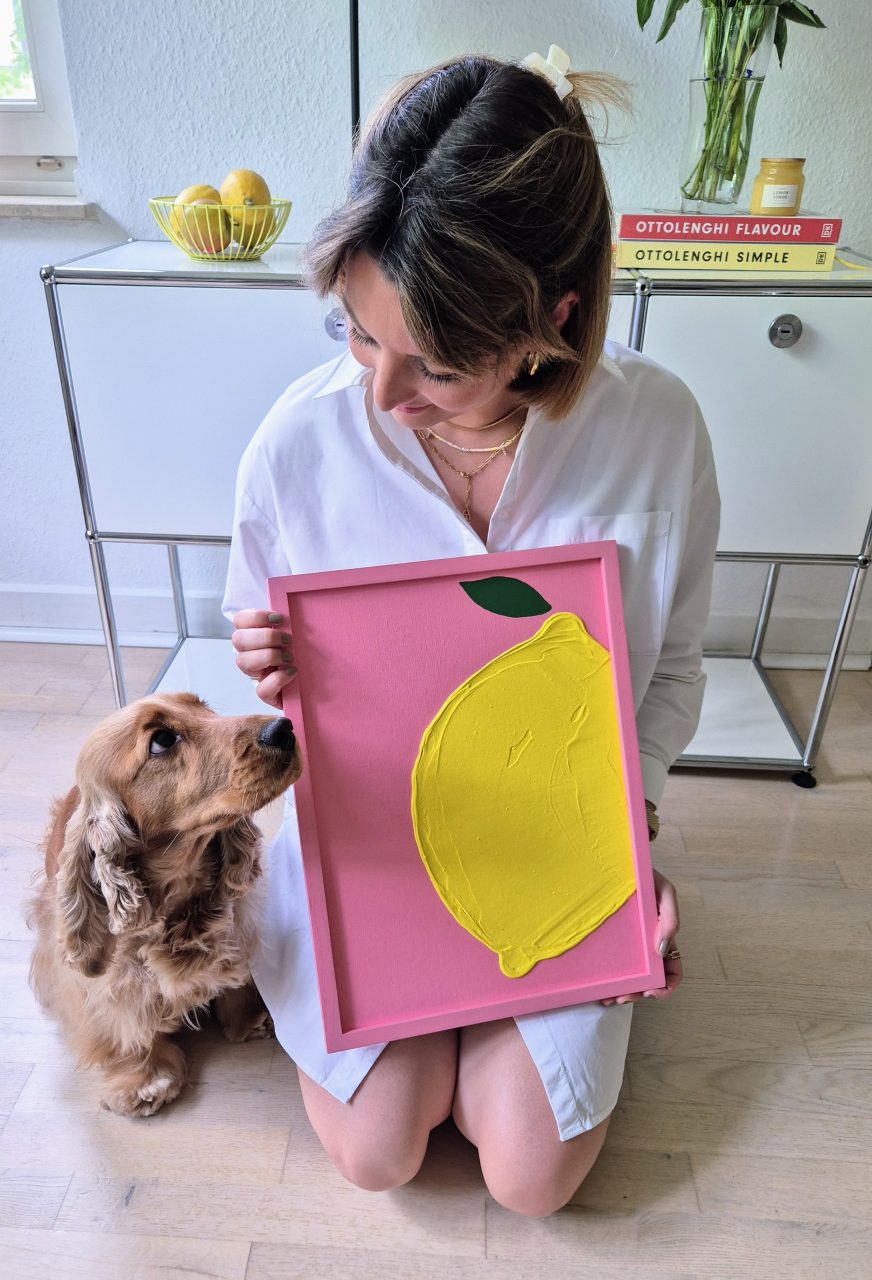 Strukturbild mit Zitronenmotiv auf pinkem Hintergrund. Gehalten von einer jungen Frau und der Hund (Cocker Spaniel) sitzt daneben.