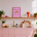 Moderne Küche in hellrose im Scandi Style. Darüber ein Holzregal mit einem Kunstwerk gestreifter Hintergrund in Weiß und Pink mit einem Croissant aus Strukturpaste und der Aufschrift "AYES SAINT CROISSANT".