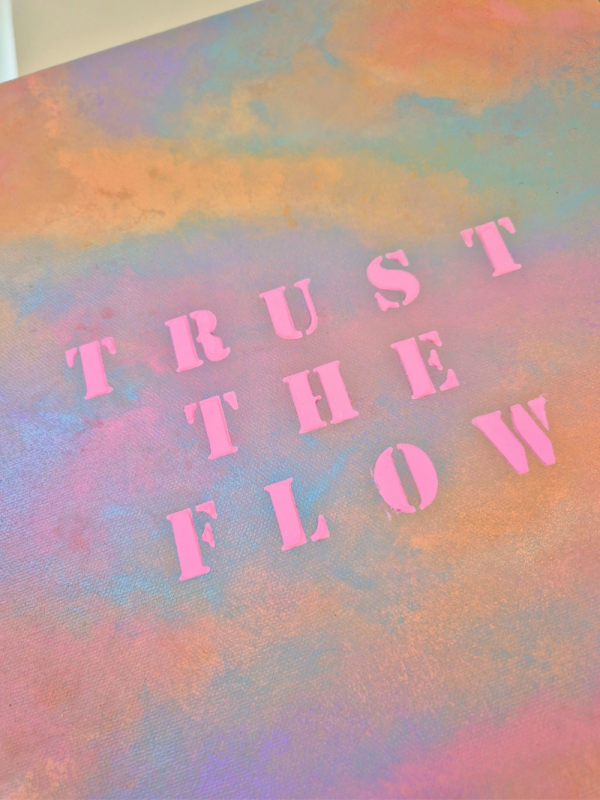 Nahaufnahme abstraktes Kunstwerk mit pastellfarbenem Himmel in Rosa, Blau und Violett sowie dem Schriftzug „TRUST THE FLOW“ in leuchtend pinken Buchstaben aus Strukturpaste.
