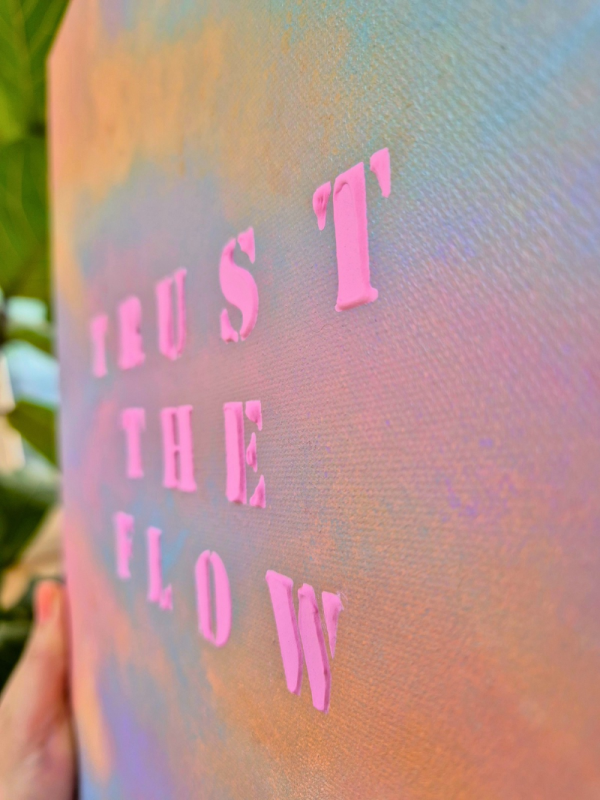 Nahaufnahme seitlich abstraktes Kunstwerk mit pastellfarbenem Himmel in Rosa, Blau und Violett sowie dem Schriftzug „TRUST THE FLOW“ in leuchtend pinken Buchstaben aus Strukturpaste.