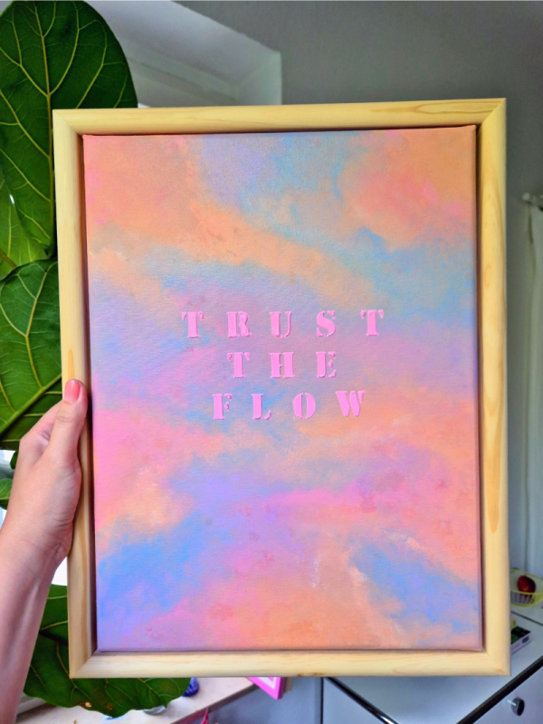 Nahaufnahme frontal abstraktes Kunstwerk mit pastellfarbenem Himmel in Rosa, Blau und Violett sowie dem Schriftzug „TRUST THE FLOW“ in leuchtend pinken Buchstaben aus Strukturpaste.